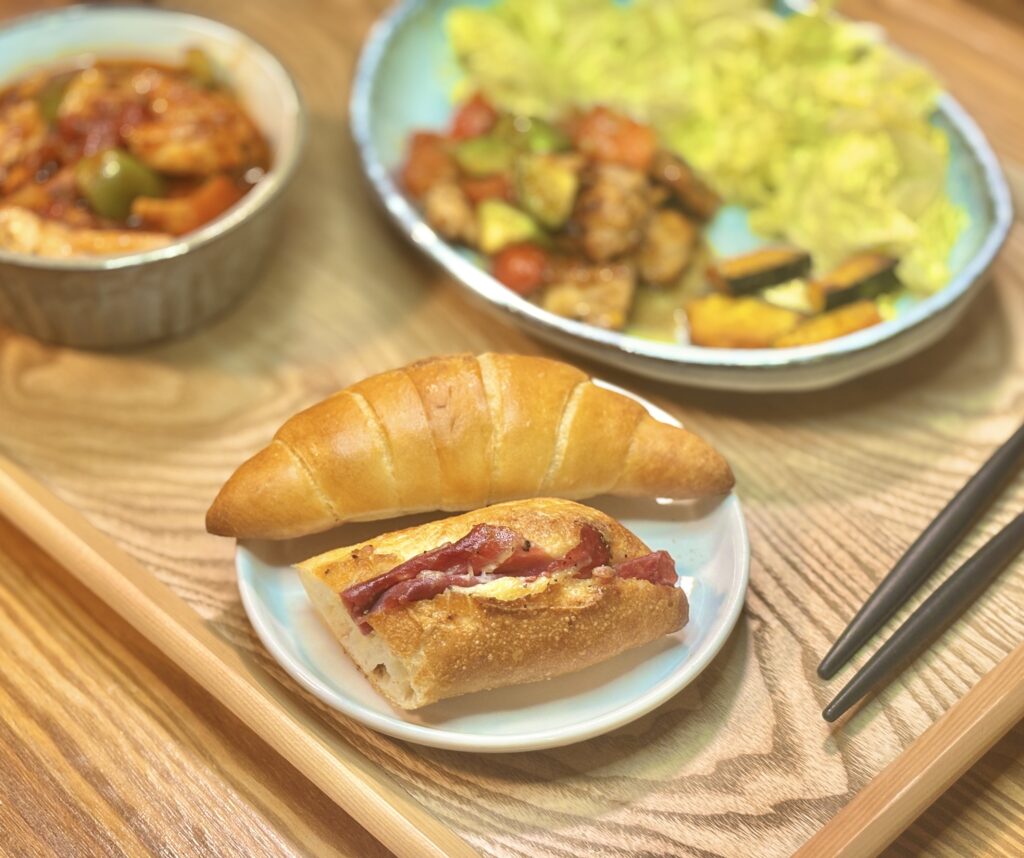 小皿にイスズベーカリーのパン2種。つやのあるロールパンと、ベーコン入りのハード系パンが手前に置かれ、奥におかずがぼんやり写る。