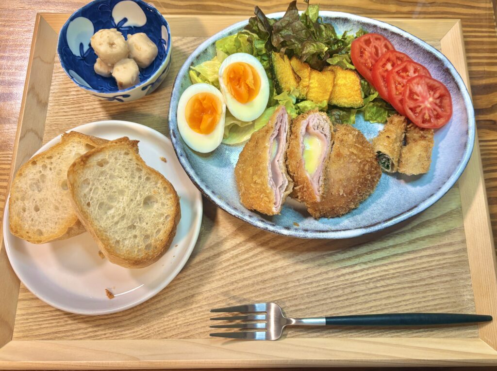 木製トレーの上に並べられたワンプレートごはん。青い楕円皿には、中央に切り分けた豚ロース肉のコルドンブルーと豚ロースの紫蘇巻き揚げが盛られ、まわりにトマトスライス、かぼちゃの素揚げ、ゆで卵、葉野菜のサラダが彩りよく添えられている。左には白い小皿にバゲット2切れ、上には青い小鉢に里芋煮があり、手前にフォークが置かれている。