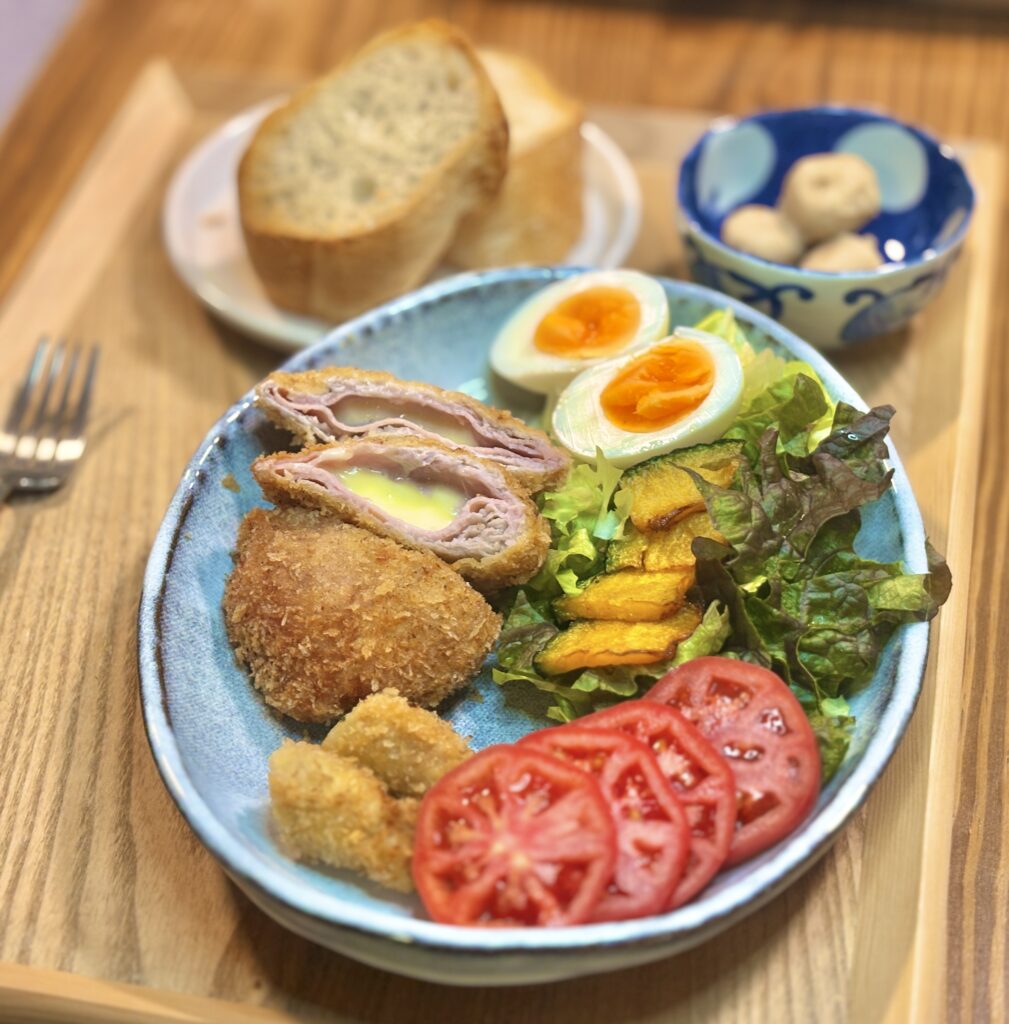 青い楕円皿を斜め上から撮影した食卓写真。手前には衣がこんがり揚がった豚ロース肉のコルドンブルーがあり、半分に切った断面からハムととろけるチーズが見えている。横には豚ロースの紫蘇巻き揚げ、トマトスライス、かぼちゃの素揚げ、ゆで卵、葉野菜のサラダが並び、背景にバゲットと里芋煮の小鉢がやわらかく写っている。