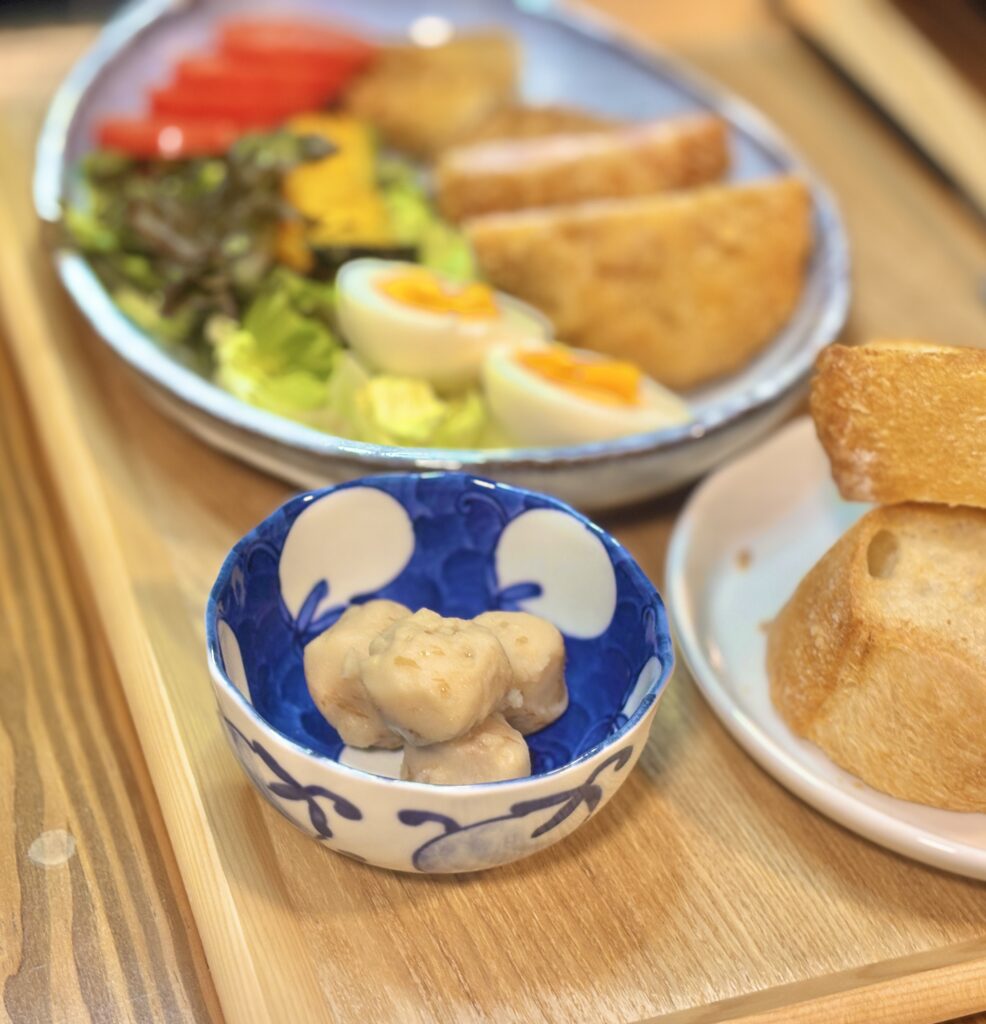 青と白の模様の小鉢に盛られた里芋煮を主役にした写真。やわらかく煮含めた里芋が数個入っており、後ろにはぼかしてワンプレートのおかずとゆで卵、トマト、バゲットが見える。木のトレーと器の色合いがあたたかく、やさしい食卓の雰囲気が伝わる一枚。