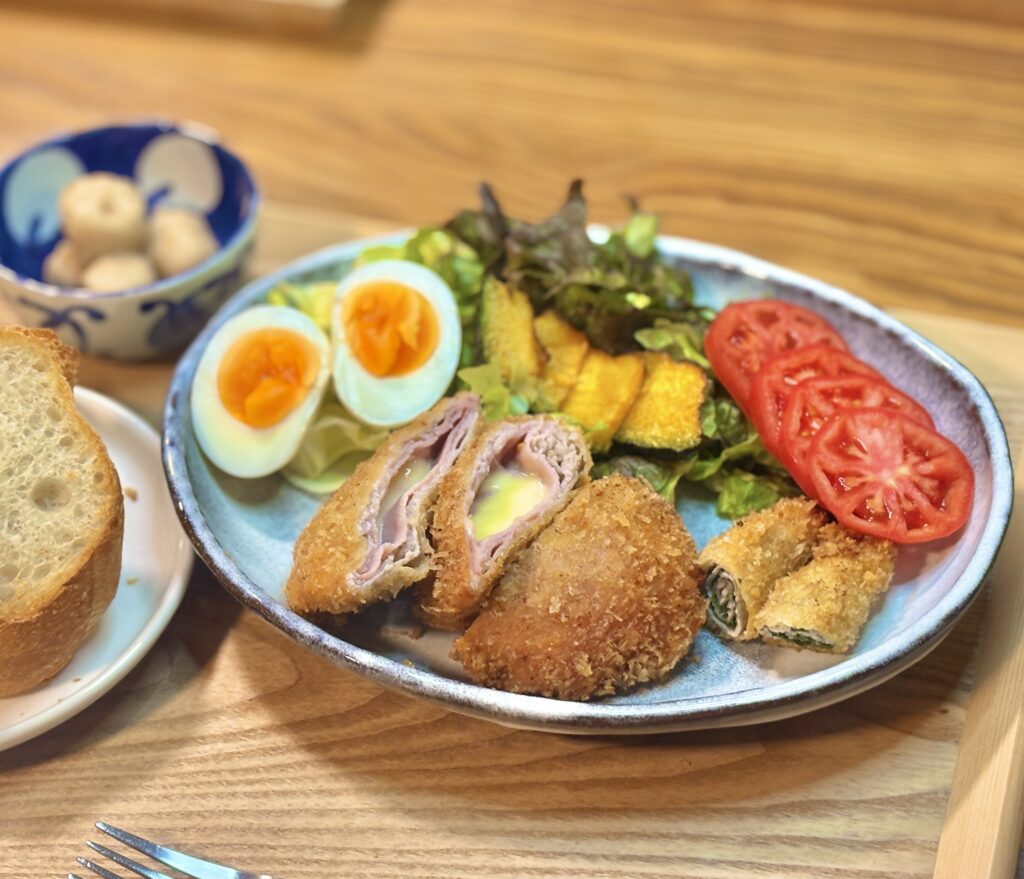 メインのおかずを中心に捉えたワンプレート写真。青い楕円皿の上に、半分に切って中のハムとチーズが見える豚ロース肉のコルドンブルーと、豚ロースの紫蘇巻き揚げが並ぶ。まわりにはトマトスライス、かぼちゃの素揚げ、半熟気味のゆで卵、葉野菜のサラダが彩りよく添えられ、左端にバゲット、奥に里芋煮の小鉢が少し写っている。