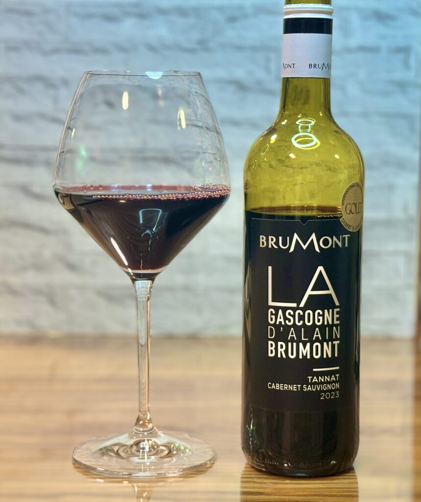 木目のテーブルの上に置かれた赤ワインのボトルとワイングラスの写真。グラスには深い赤色のワインが注がれ、隣には「BRUMONT LA GASCOGNE D’ALAIN BRUMONT TANNAT CABERNET SAUVIGNON 2023」と書かれたボトルが立っている。食事に合わせる一杯として、落ち着いた大人の雰囲気が感じられる。