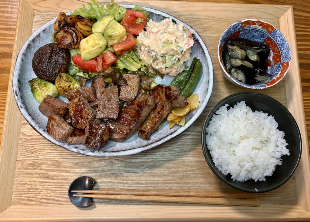 木製トレーの上に並べられた黒毛和牛のサイコロステーキとステーキ、ポテトサラダ、アボカドとトマトのサラダ、ナスの揚げ浸し、ごはんの夕食膳。焼き色のついた牛肉に、彩り豊かな副菜が添えられた満足感のある和洋折衷の一皿。