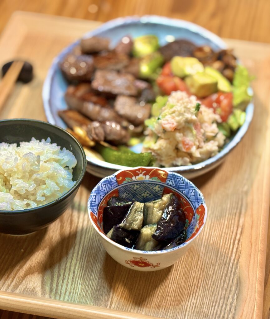 手前にナスの揚げ浸しの小鉢と白ごはん、奥にぼんやりと黒毛和牛のステーキプレートが写る夕食写真。主菜と副菜を立体的に切り取った構図で、やわらかな光の中に家庭料理のやさしさが感じられる一枚。