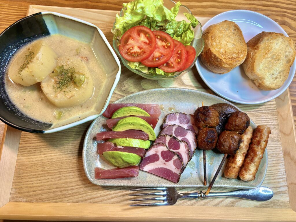 鳥つくね串、ソーセージリンクス、パストラミポーク、アボカドとキハダマグロの盛り合わせに、コンソメで煮た洋風大根煮、トマトとレタスのサラダ、焼きバゲットを添えた洋風ワンプレートディナーの全体写真。