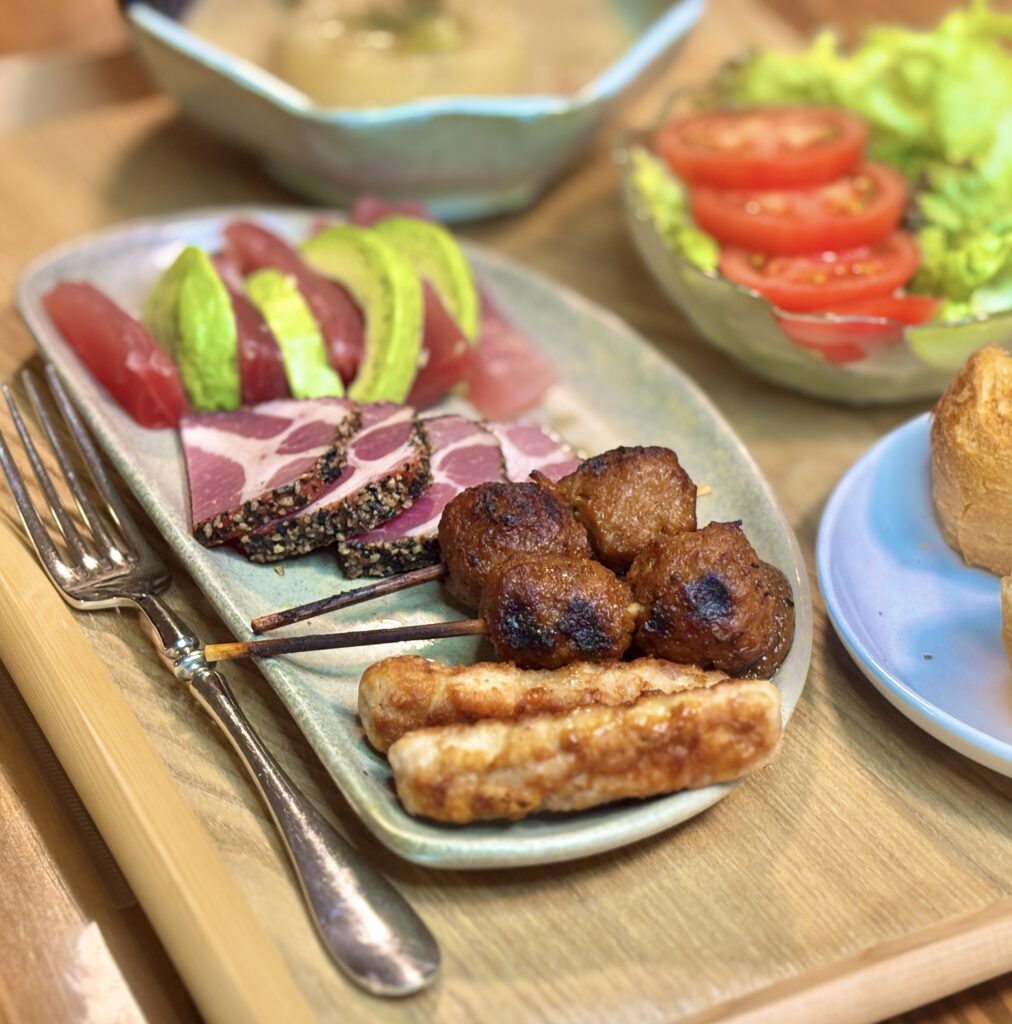 鳥つくね串、ソーセージリンクス、パストラミポーク、アボカドとキハダマグロを盛り合わせた洋風おつまみプレート。奥に洋風大根煮とトマトサラダが並ぶ家庭の食卓。