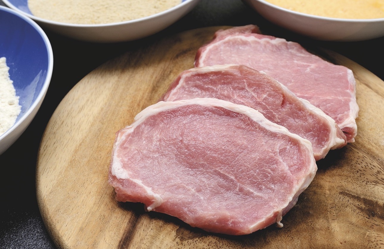 豚ロース肉の厚切りが3枚、木製のカッティングボードの上に並べられている様子。周囲にはパン粉や小麦粉などの下ごしらえ用の材料が入った器が置かれており、トンテキやとんかつの調理前の準備シーンを写した一枚。