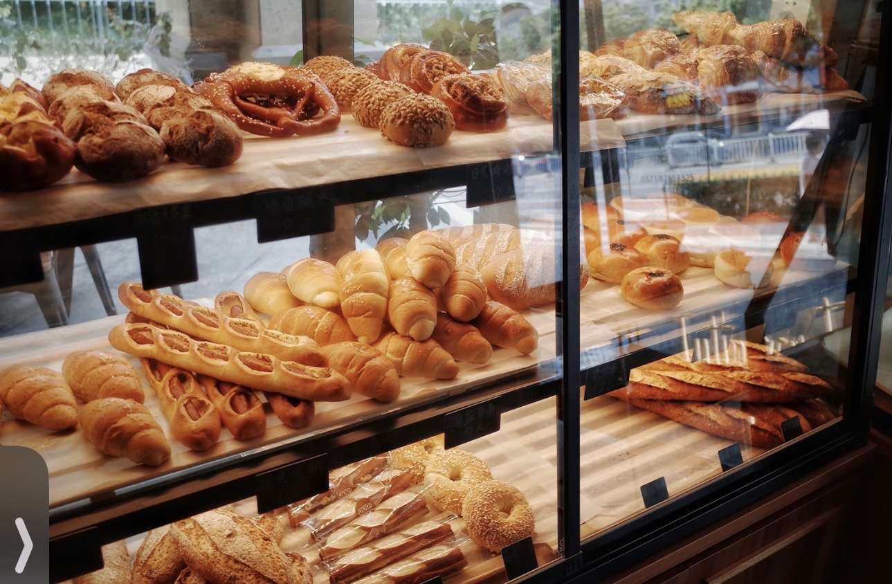 ガラスケースの中に並ぶ焼きたてのパン。クロワッサン、ソーセージ入りパン、プレッツェル、バゲットなどさまざまな種類が木の棚に整然と並び、ベーカリー店内の温かい雰囲気と外の街並みの映り込みが見えるショーケースの風景。