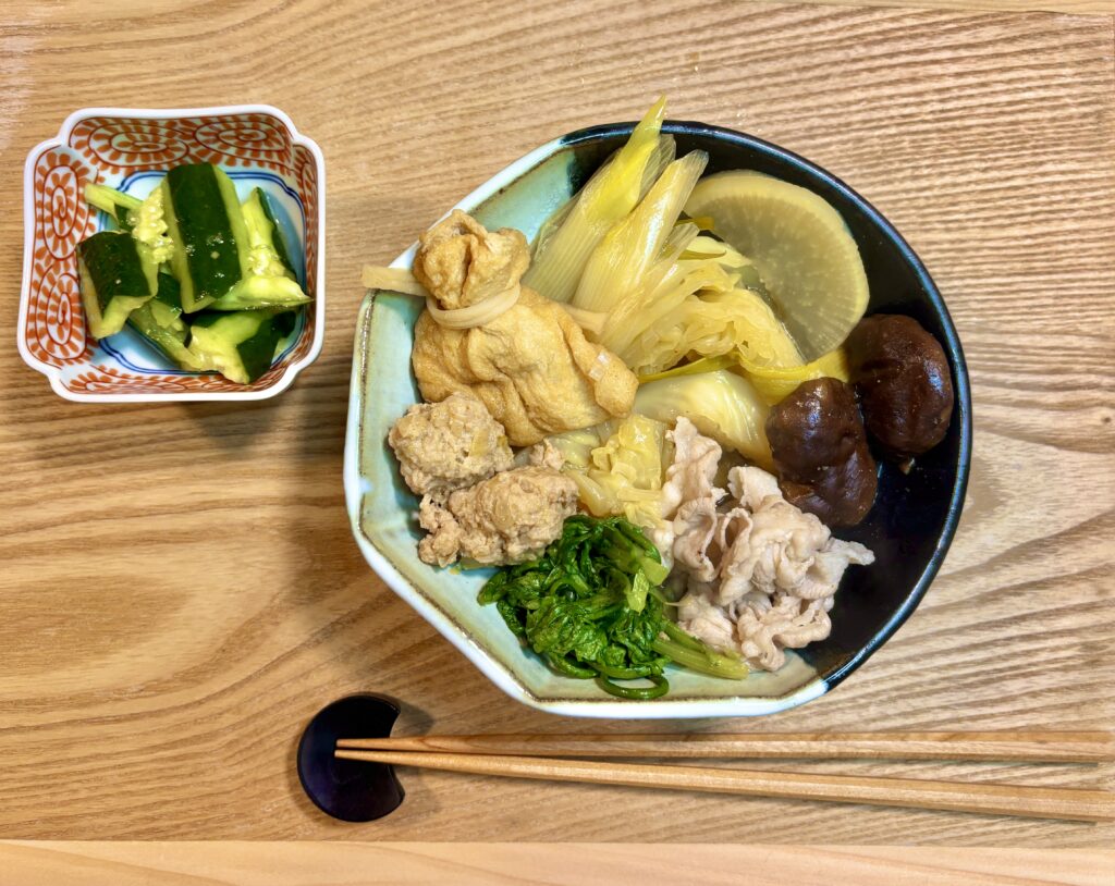ラーメン来来亭のスープで作った鍋料理を上から撮影した写真。器には白菜、白ねぎ、豚肉、鶏つくね、しいたけ、青菜、巾着、大根が彩りよく盛られ、横には小鉢に入ったきゅうりのたたきが添えられている。木のテーブルに箸と箸置きも並び、あたたかいおうち鍋の雰囲気が伝わる一枚。