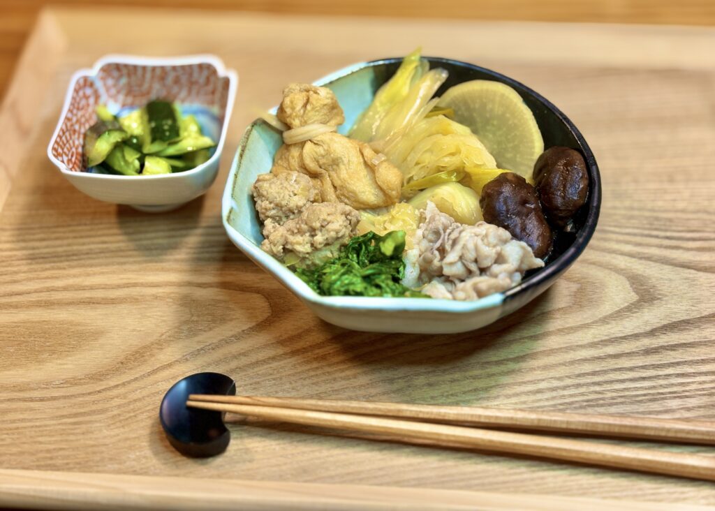 ラーメン来来亭のスープで作った鍋料理を斜め前から写した写真。器の中には白菜、白ねぎ、豚肉、鶏つくね、しいたけ、青菜、巾着、大根がたっぷり入り、手前には箸、奥にはきゅうりのたたきの小鉢がぼんやり映っている。やさしい塩味の鍋らしい、ほっとする食卓の雰囲気が感じられる。