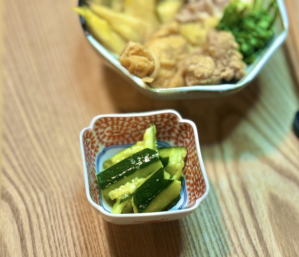 小鉢に盛り付けたきゅうりのたたきのアップ写真。つやのある緑色のきゅうりが食べやすい大きさに切られて入り、背景にはラーメン来来亭のスープで作った鍋料理がやわらかくぼけて見える。さっぱりした副菜として鍋に添えた様子が伝わる一枚。