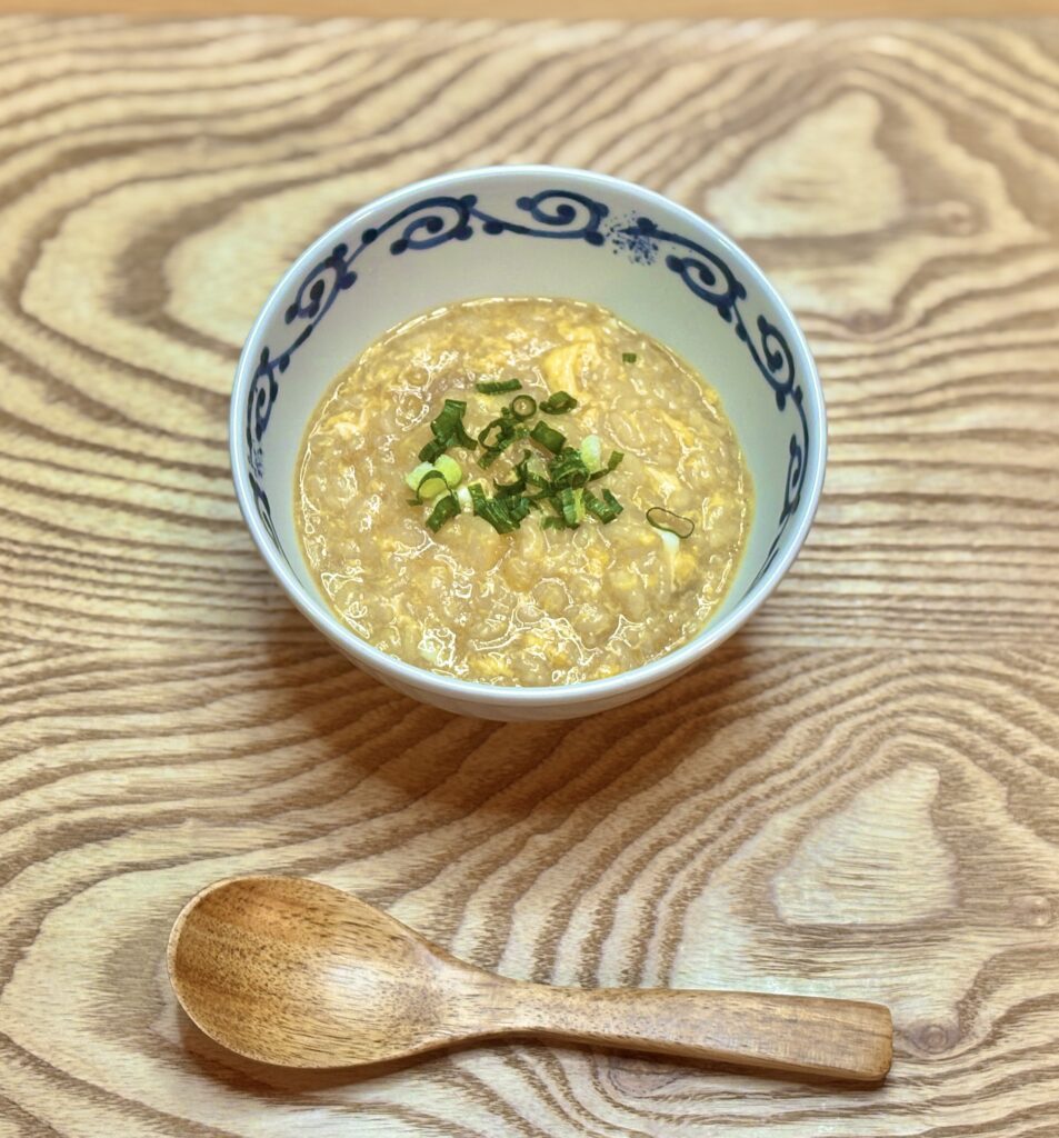 ラーメン来来亭のスープで作った鍋の締めのおじやを写した写真。器には鍋の旨味が溶け込んだとろりとした雑炊が盛られ、卵のふんわりした食感と青ねぎの彩りが引き立っている。手前には木のスプーンが置かれ、鍋の最後までおいしく楽しめる一品であることが伝わる。