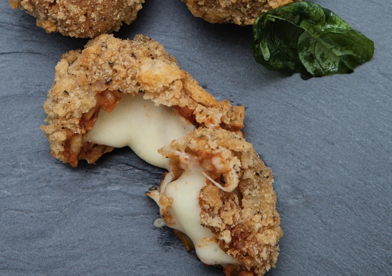 半分に切られた豚ロース肉のコルドンブルーをアップで撮影した写真。サクサクの衣の中からハムととろけたチーズがあふれ出し、断面からチーズがやわらかく伸びている。黒いプレートの上に置かれ、横には彩りとして緑の葉が添えられている。