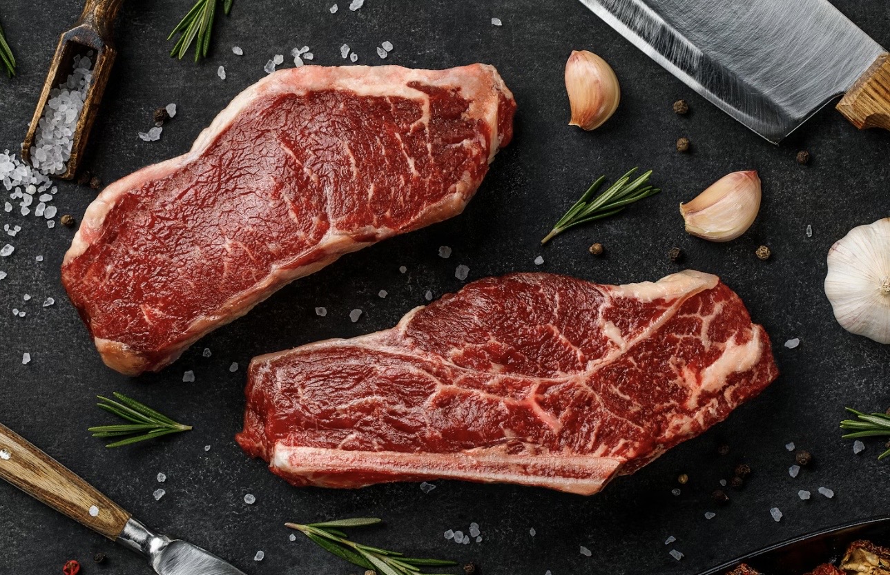 生の牛ステーキ肉が2枚、黒い石のような背景の上に並べられている。周囲には粗塩、黒胡椒、にんにく、ローズマリーが散らされ、両側には木製ハンドルのナイフが置かれており、調理前のステーキ素材をスタイリッシュに演出した食材イメージ写真。