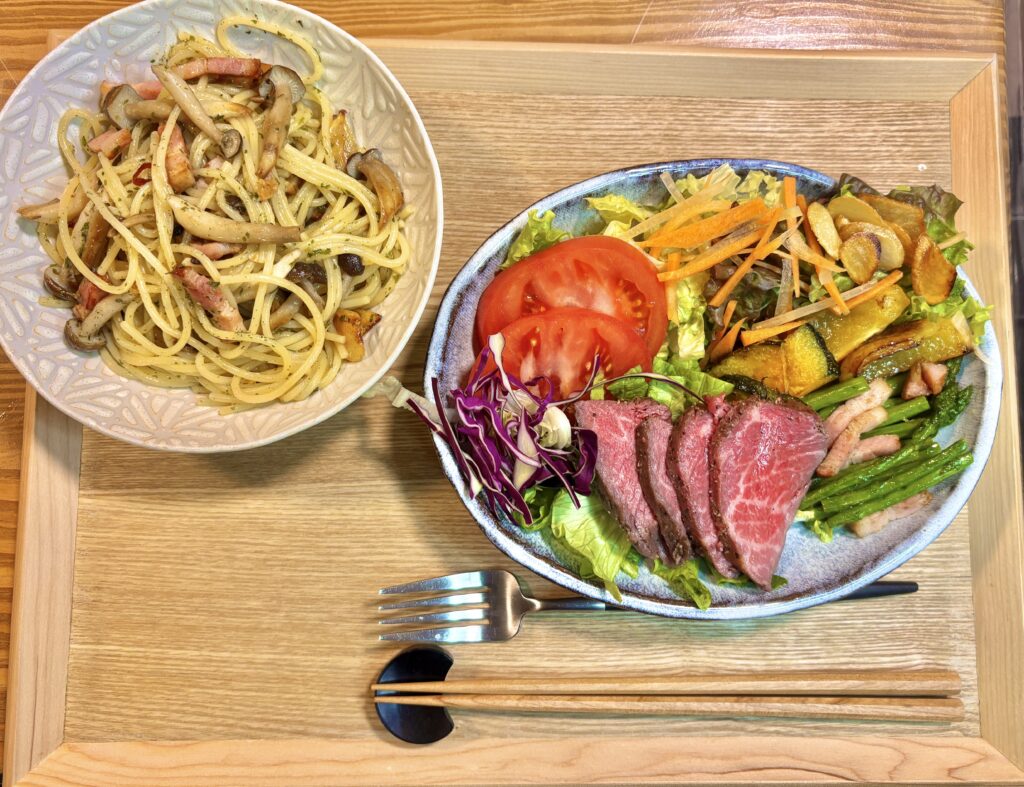 木製トレーの上に並べられた晩ごはんプレート。ベーコンとしめじのぺぺロンチーノのパスタ皿と、ローストビーフを中心に彩り野菜、焼きかぼちゃ、ベーコンとアスパラのガーリック炒めが盛られたサラダプレートが美しく配置された旦那キッチンのごちそうディナー。

