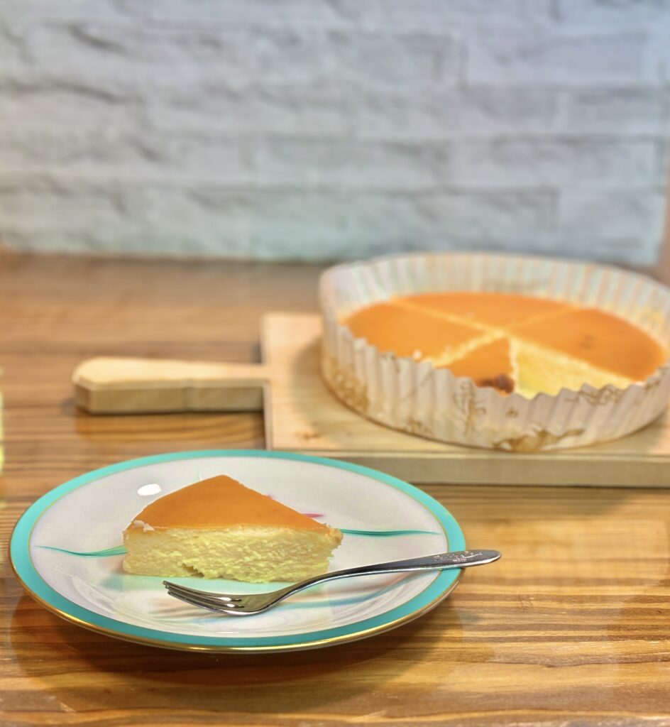 皿に盛り付けた御用邸チーズケーキのカットと、後ろにホールのチーズケーキ。しっとり濃厚な那須高原チーズガーデンの人気スイーツ。