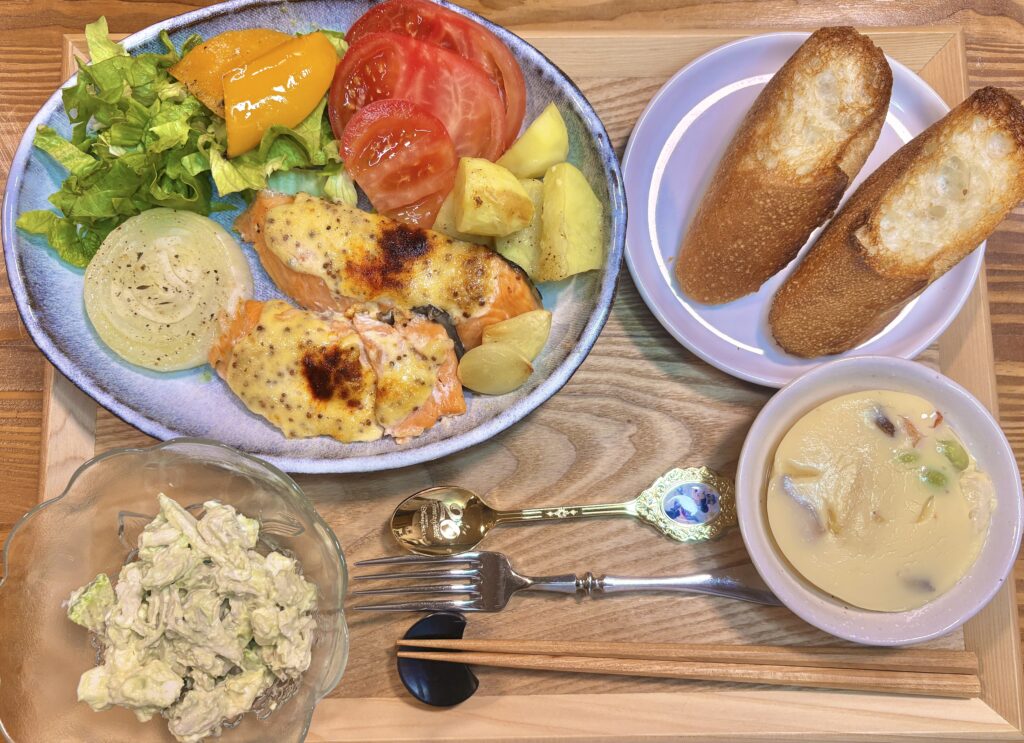 木製トレーの上に並べた晩ごはん全体の写真。オーブンで焼いた甘塩シャケのガーリックマスタードマヨネーズがメインで、レタス、トマト、パプリカ、じゃがいも、玉ねぎを添えたワンプレートに、市販の茶碗蒸し、アボカドとクリームチーズと鶏胸肉のワサビマヨネーズ和え、サンノトーレのバゲットを合わせた食卓。