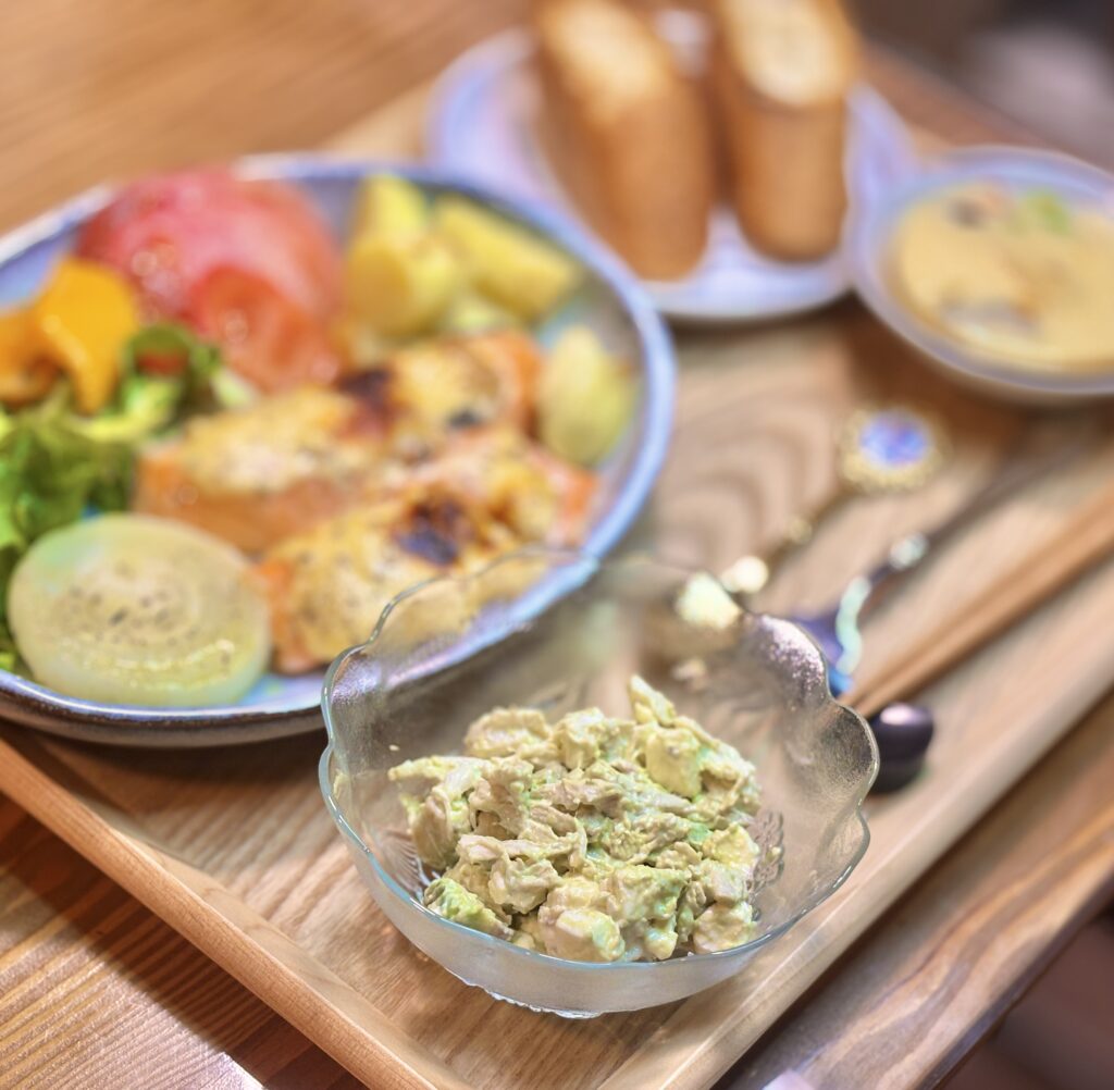 ガラスの器に盛り付けたアボカドとクリームチーズと鶏胸肉のワサビマヨネーズ和えを手前に写した写真。奥には少しぼかして、シャケのオーブン焼き、茶碗蒸し、バゲットが並び、クリーミーな副菜のおしゃれな存在感が引き立つ一枚。