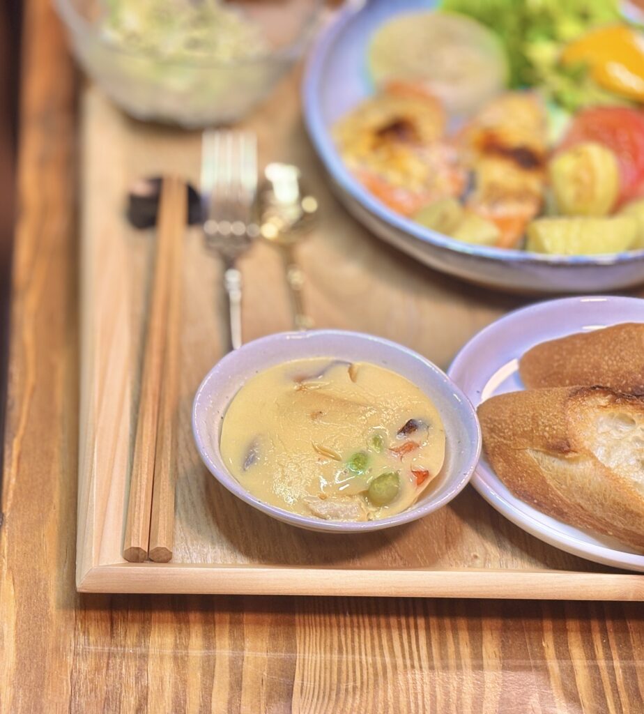 市販の茶碗蒸しを小さな器に入れて食卓に添えた写真。なめらかな表面の中に具材が見え、横には焼いたバゲット、奥にはシャケのガーリックマスタードマヨネーズ焼きがぼけて写る、手軽さとやさしい味わいが伝わる一枚。