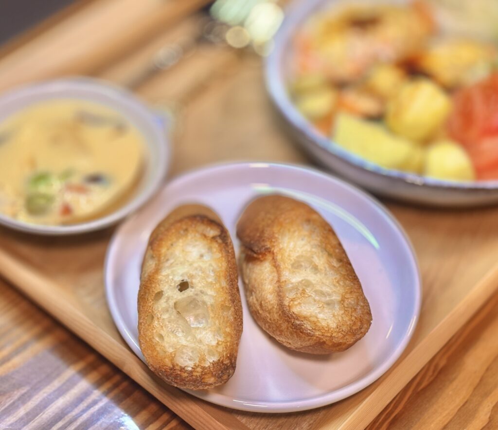 こんがり焼いたサンノトーレのバゲットを2切れ、小さなピンクの皿にのせた写真。外は香ばしく中はふんわりした焼き色が美味しそうで、奥には茶碗蒸しやシャケのオーブン焼きがぼんやり写り、食卓の温かい雰囲気が感じられる。