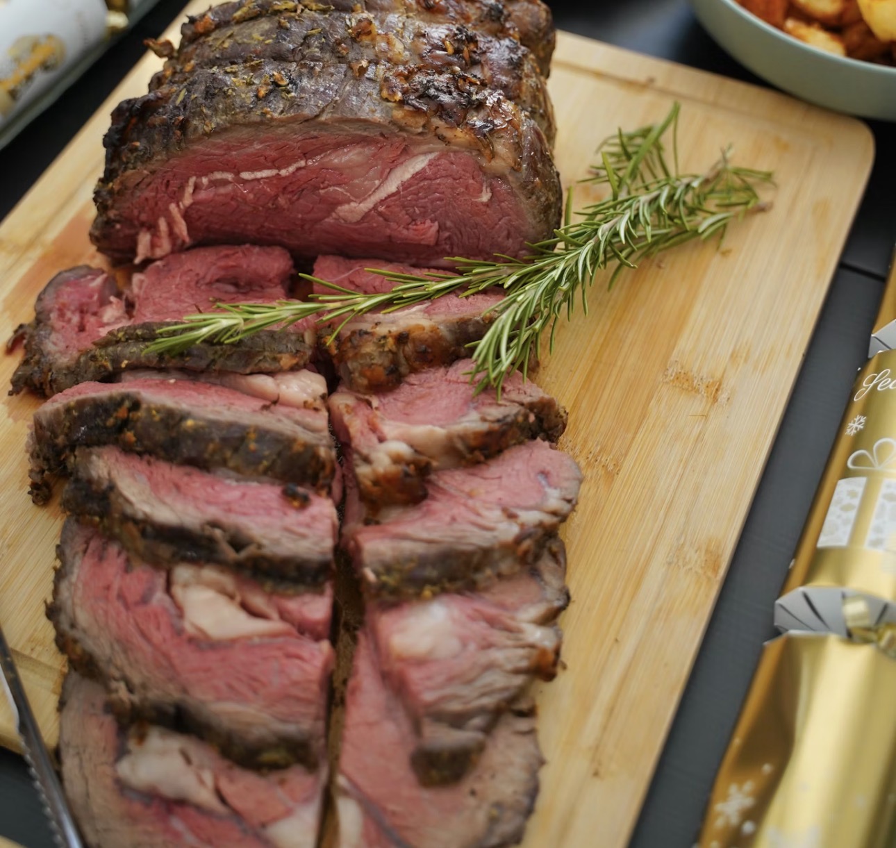 木製カッティングボードの上に並べられたローストビーフ。外側は香ばしく焼き色がつき、中は美しいロゼ色のレアに仕上がっており、スライスされた牛肉の断面からジューシーな肉汁が感じられる。仕上げにローズマリーが添えられたごちそう感のある一皿。