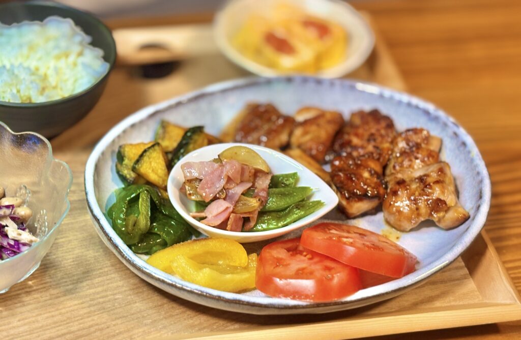 ハニーマスタードチキンと焼き野菜、スナックえんどうとベーコンのガーリック炒め、トマトを盛り付けた旦那キッチンの晩ごはんプレート。ご飯と明太子卵焼きが添えられた家庭料理の食卓。