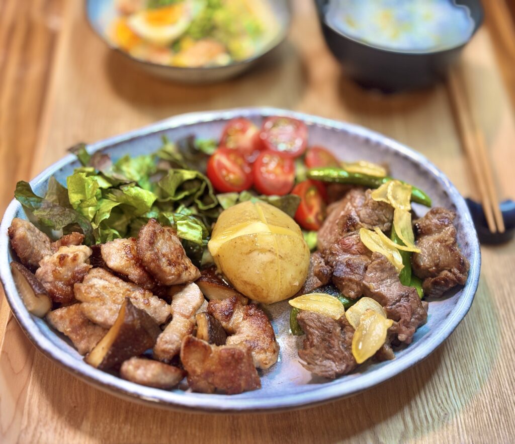 ガーリックチップをのせたサイコロステーキとスナックえんどう、トンテキ椎茸焼き、中央にバターが溶けるじゃがバターとミニトマトサラダの盛り合わせ。