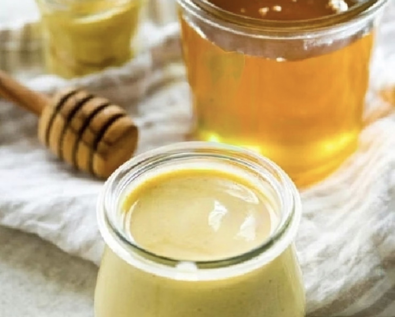 はちみつと粒マスタードを使ったハニーマスタードソースのイメージ写真。ガラス瓶に入った黄金色のはちみつと木製ハニーディッパー、手前にはクリーミーなハニーマスタードソースが入った小瓶が並ぶ料理素材写真。