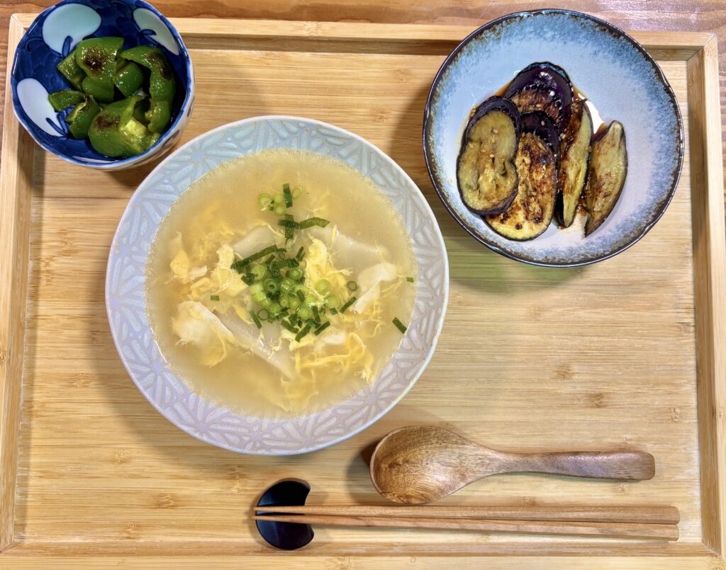 水餃子入り卵スープとナスのごま油炒め、ピーマンの焼きびたしを並べた和風献立の俯瞰写真。木製トレーに盛り付けたシンプルで体にやさしい夜ごはん