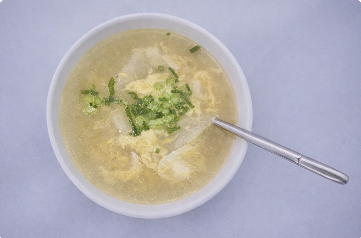 水餃子入り卵スープの俯瞰写真。鶏ガラスープと帆立の旨みを合わせた黄金色のスープに、ふんわり卵と青ねぎをトッピングし、スプーンを添えたシンプルでやさしい夜食メニュー