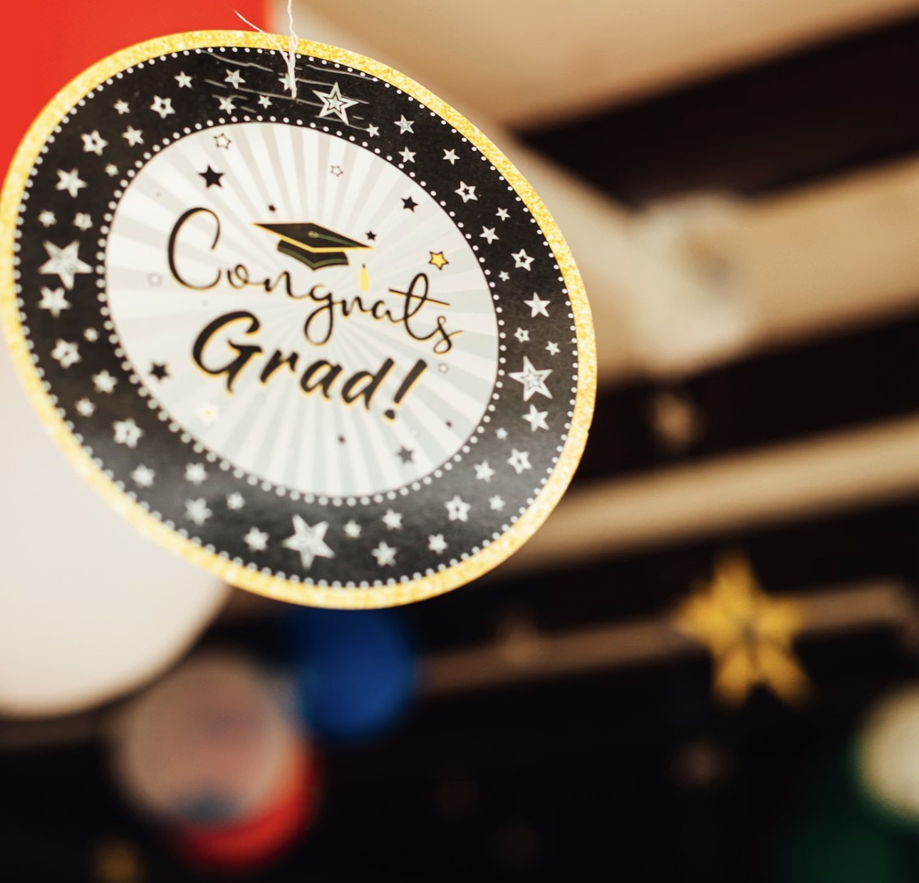 卒業祝いの「Congrats Grad!」と書かれた丸型デコレーションのクローズアップ。黒とゴールドを基調にした華やかなデザインで、背景には卒業帽や星の飾りがぼかして写っている。
