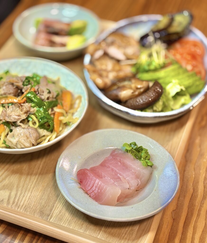 桜ビンチョウマグロのお造りをシンプルに盛り付けた一皿と、焼きそばやおつまみを並べた家飲みディナーの全体風景
