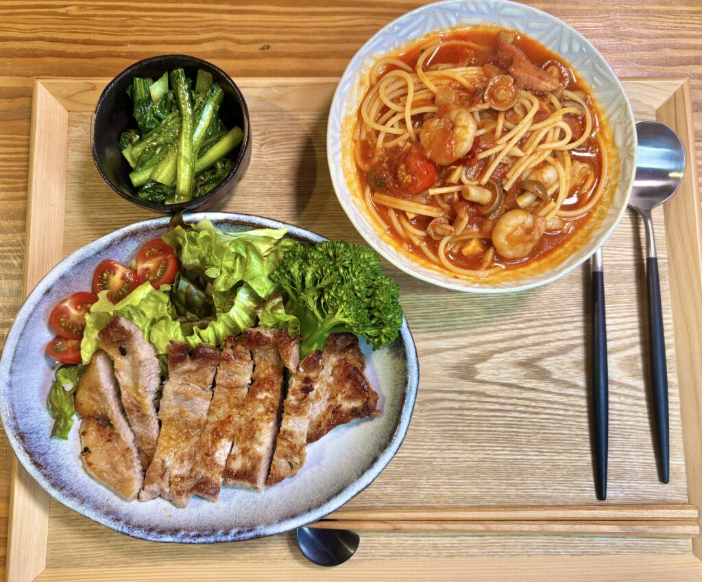 旦那キッチン特製、岩塩と小麦粉で外はカリッと中はジューシーに焼き上げたトンテキ。付け合わせのブロッコリーとトマト、小松菜のガーリック炒め、奥にはトマトジュースで作る時短スープパスタが並んだボリューム満点の夕食献立風景。