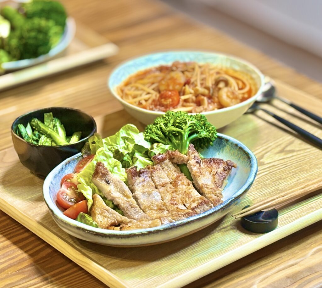 旦那キッチン特製、岩塩と小麦粉で外はカリッと中はジューシーに焼き上げたトンテキ。付け合わせのブロッコリーとトマト、小松菜のガーリック炒め、奥にはトマトジュースで作る時短スープパスタが並んだボリューム満点の夕食献立風景。