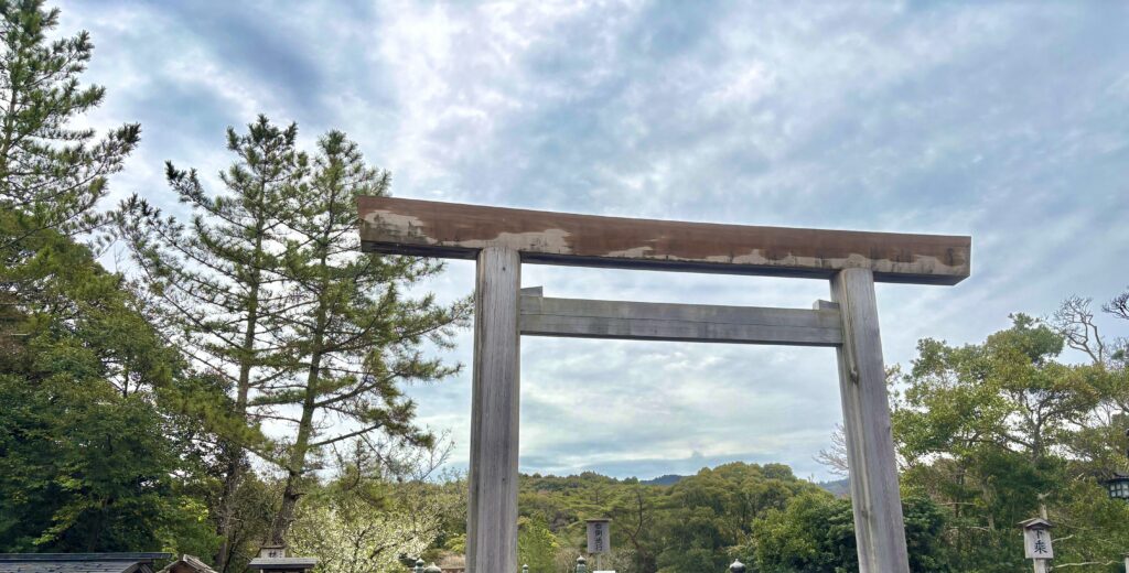 三重観光で訪れた雨上がりの伊勢神宮。豊かな緑の木々と曇り空を背景にそびえ立つ、神聖で立派な木造の大鳥居の風景