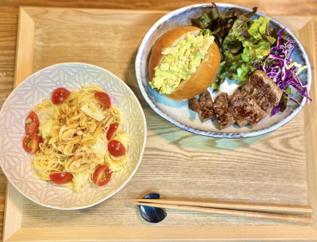 木製トレーの上に並べられた夕食プレート。春キャベツとサクラエビのガーリック冷製パスタ、アボカドとクリームチーズと鶏胸肉のマヨネーズごまドレ和えサンド、牛モモ肉のソテー、彩りサラダがバランスよく配置された食卓の様子。