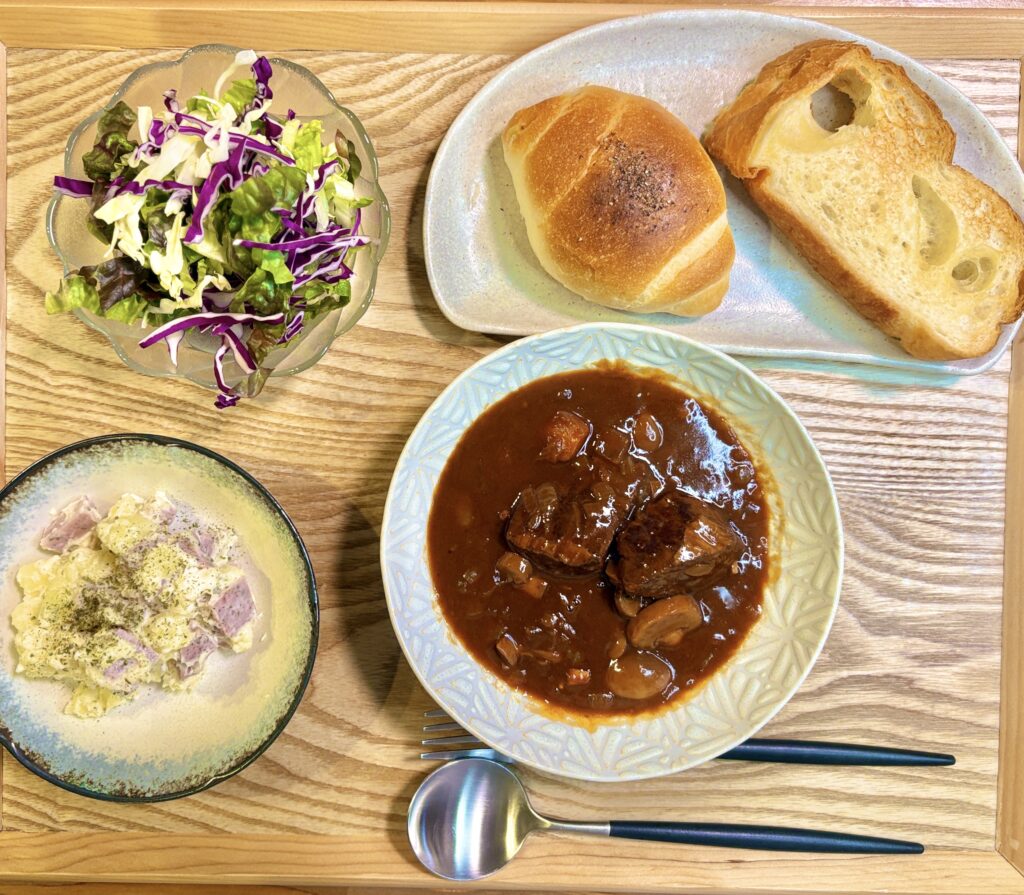 木製トレーの上に並んだ手作りディナーセット。和牛もも肉のビーフシチューを中心に、明宝パセリソーセージと新じゃが・クリームチーズのポテトサラダ、彩り野菜サラダ、塩パンと塩バターフランスを添えた洋食プレート。

