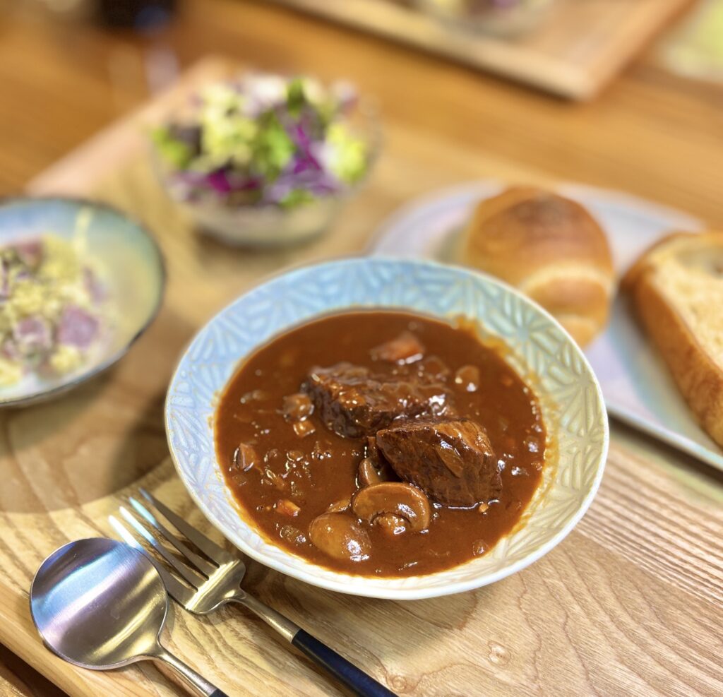 とろみのある濃厚ソースに大きめの和牛もも肉とマッシュルームが入ったビーフシチュー。スプーンとフォークを添えた温かみのあるテーブルコーディネート。
