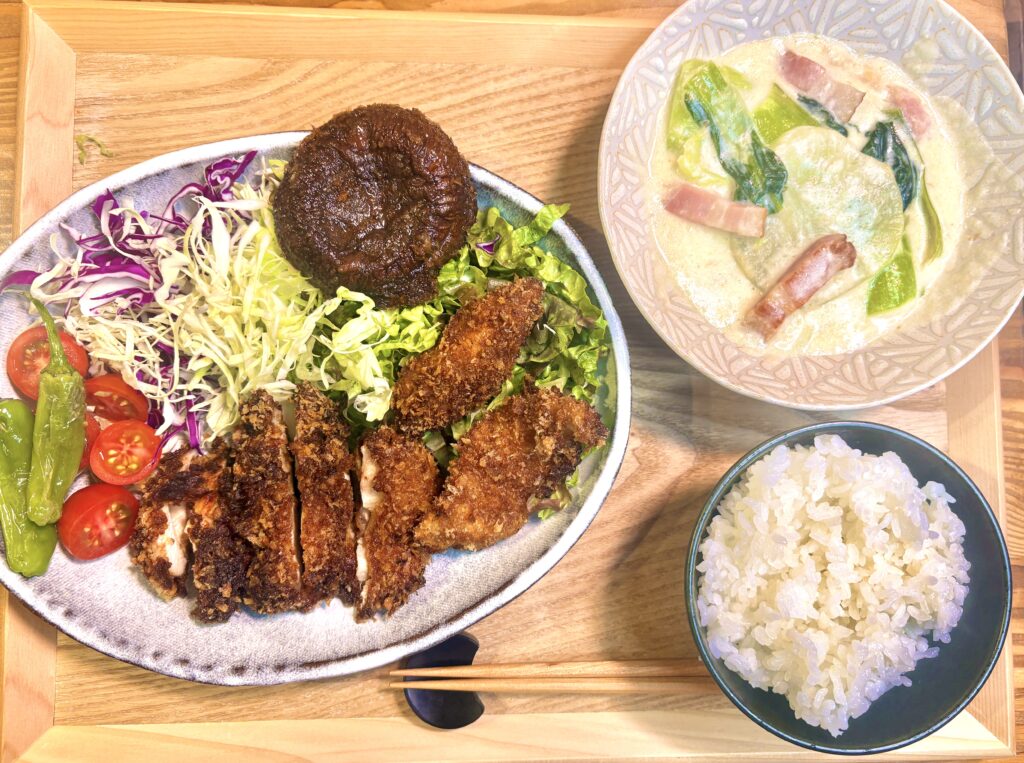 木製トレーの上に並べられた晩ごはんの全体写真。大皿には、いわむらさんのレシピを参考に作った悪魔のチキンカツが盛り付けられており、胸肉ともも肉の両方を使った食べ比べが楽しめる一皿になっている。こんがりきつね色に揚がったチキンカツのほか、焼きししとう、半分にカットしたミニトマト、千切りキャベツ、紫キャベツ、レタス、焼き椎茸が彩りよく添えられていて、見た目にも華やか。別皿には青梗菜と大根とベーコンのクリーム煮がたっぷり盛られ、ごはん茶碗と箸も並んだ、家庭的で満足感のある定食スタイルの夕食風景を写した料理写真。