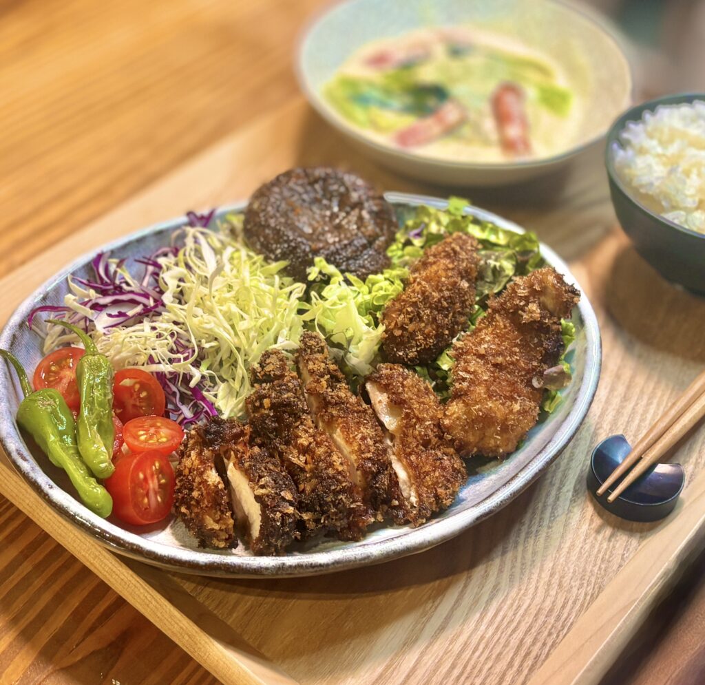 斜め前から撮影した食卓の料理写真。主役の悪魔のチキンカツは、胸肉ともも肉を使って作られており、衣はサクサク、中はやわらかそうな断面までしっかり見えるようにカットされている。付け合わせには焼きししとう、ミニトマト、千切りキャベツ、紫キャベツ、レタス、焼き椎茸がたっぷり添えられ、ボリュームのあるワンプレートごはんとして仕上がっている。背景には青梗菜と大根とベーコンのクリーム煮、ごはん茶碗がやさしくぼけて写り、手前のチキンカツの香ばしさと揚げたて感がより引き立つ構図。手作り晩ごはんや家庭料理ブログ、チキンカツ献立の記事にぴったりの一枚。