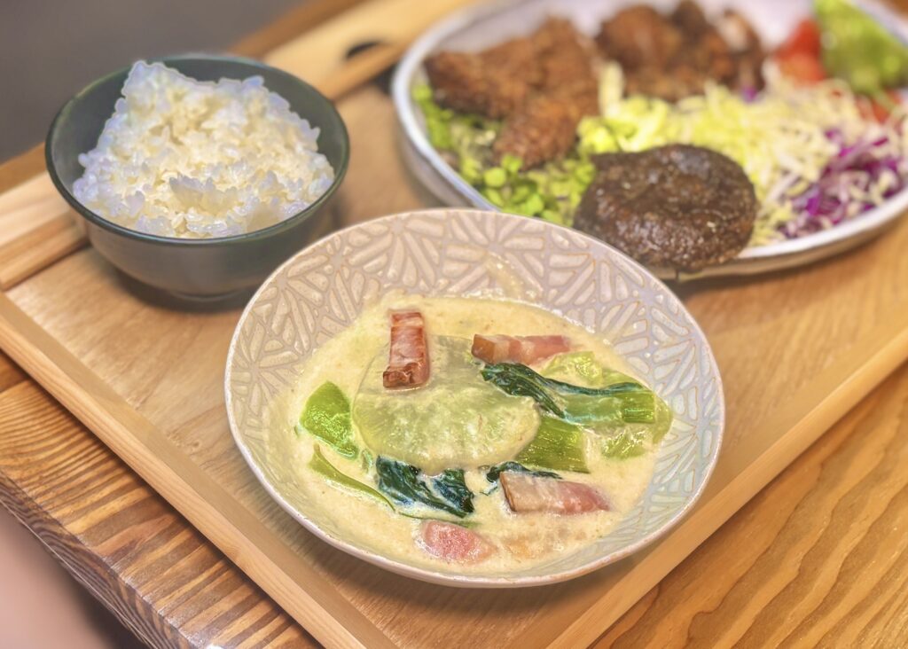 斜め前から撮影した食卓の料理写真。主役の悪魔のチキンカツは、胸肉ともも肉を使って作られており、衣はサクサク、中はやわらかそうな断面までしっかり見えるようにカットされている。付け合わせには焼きししとう、ミニトマト、千切りキャベツ、紫キャベツ、レタス、焼き椎茸がたっぷり添えられ、ボリュームのあるワンプレートごはんとして仕上がっている。背景には青梗菜と大根とベーコンのクリーム煮、ごはん茶碗がやさしくぼけて写り、手前のチキンカツの香ばしさと揚げたて感がより引き立つ構図。手作り晩ごはんや家庭料理ブログ、チキンカツ献立の記事にぴったりの一枚。