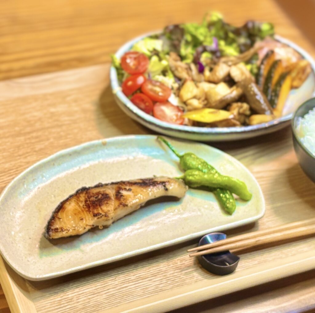 こんがり焼き目のついた銀鱈の西京焼きを中心に、ししとうを添えた和食プレート。奥には鶏肉ときのこのガリバタポン酢炒めや彩りサラダが並び、家庭で楽しむ贅沢な夕食メニューの雰囲気が伝わる一枚。
