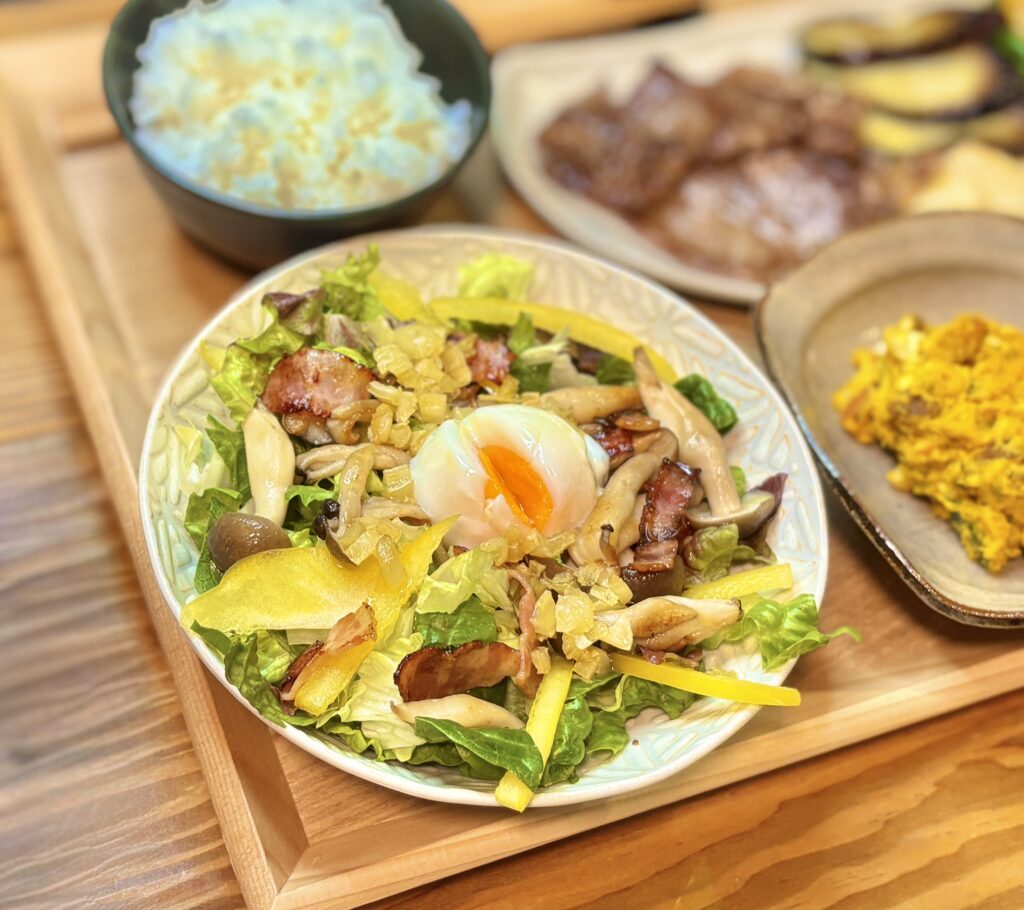 ロメインレタスの上にベーコンとしめじのガーリック白だし炒めをのせ、中央に温玉をトッピングしたシーザーサラダ。黄身が半熟でとろりとした状態。奥に和牛焼肉とかぼちゃサラダが見える。