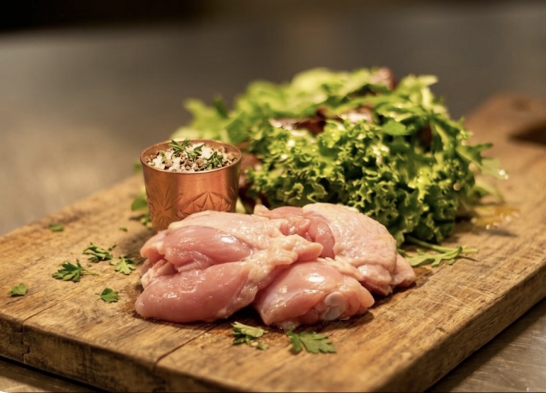 木製まな板の上に下処理された鶏もも肉と新鮮な葉物野菜、ハーブと塩が添えられた料理準備シーン、キッチンの仕込み風景