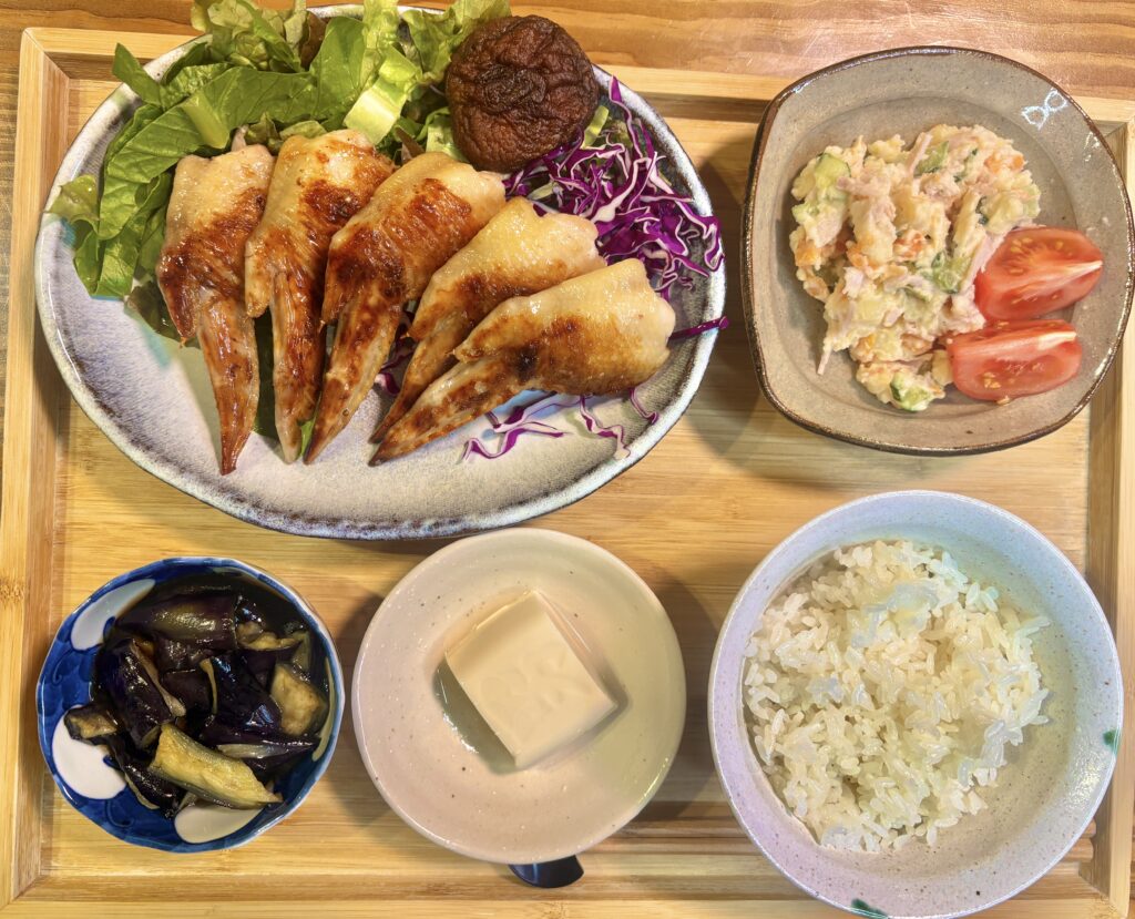 木製トレーの上に、手羽先の塩焼きをメインにした和定食風の献立が並ぶ夕食写真。こんがり焼き色のついた手羽先はレタスと紫キャベツの上に美しく盛り付けられ、付け合わせにはしいたけも添えられている。さらに、ナスの揚げ浸し、ポテトサラダ、冷奴、白ごはんが並び、家庭料理らしい温かみと栄養バランスの良さが伝わる一枚。手羽先献立、和風おかず、家庭の晩ごはん、定食スタイルの食卓写真として使いやすい画像。