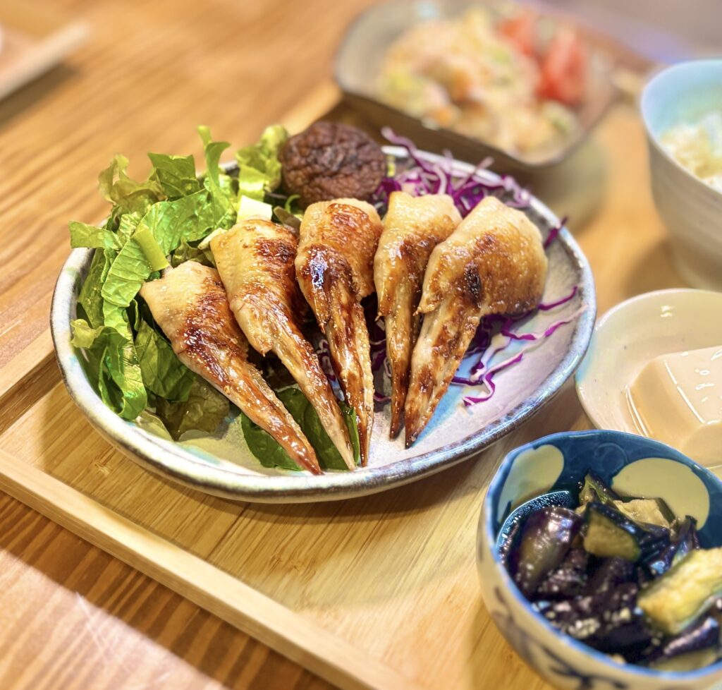 香ばしく焼き上げた手羽先の塩焼きを主役にした夕食プレートの写真。パリッと焼けた皮目が美しく、ジューシーさを感じる手羽先が5本並び、下にはレタスと紫キャベツが敷かれて彩りよく盛り付けられている。奥にはポテトサラダやごはん、手前にはナスの揚げ浸しと冷奴も見え、家庭で楽しむ手羽先メニューの魅力が伝わる構図。手羽先レシピ、手羽先の塩焼き、晩ごはんのおかず、和風献立、家庭料理のブログ用画像に適した写真。