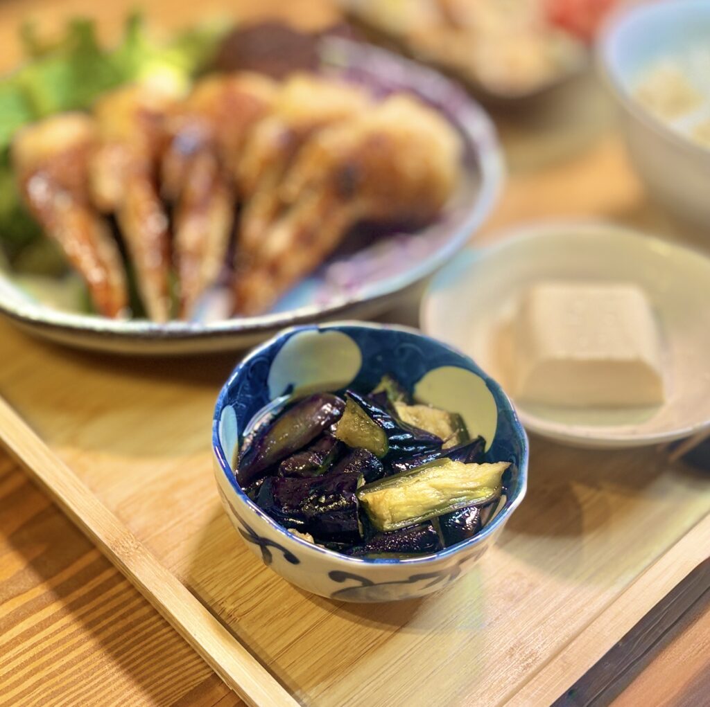 小鉢に盛り付けられたナスの揚げ浸しを主役にした和風おかずの写真。つやつやとした濃い紫色のナスにだしがしみ込み、やわらかく味わい深い仕上がりが伝わる一枚で、背景には手羽先の塩焼きや冷奴がふんわりと写り、食卓全体の雰囲気も感じられる。副菜としての上品さと、家庭料理ならではのやさしい温もりが魅力。ナスの揚げ浸しレシピ、和食の副菜、なす料理、小鉢メニュー、作り置きおかずのイメージ写真として活用しやすい画像。