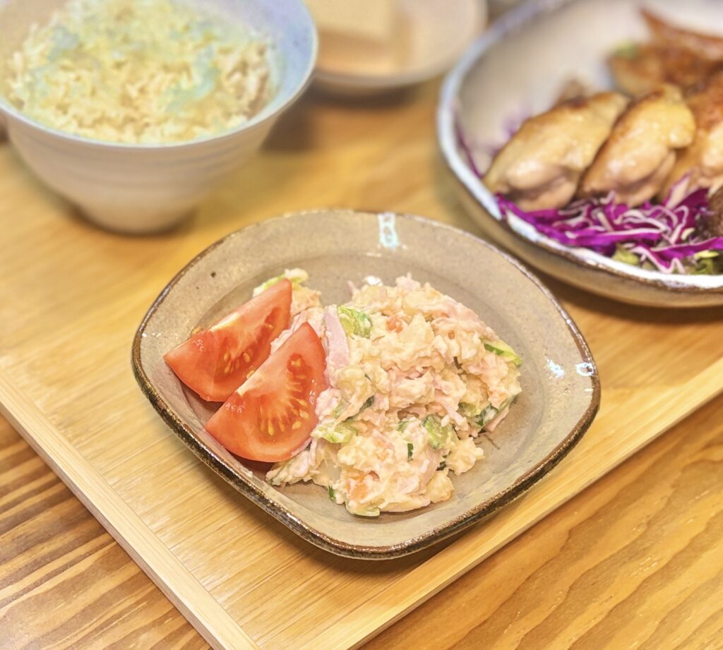 器に盛られた手作りポテトサラダとカットトマトを写した家庭料理の写真。じゃがいもにきゅうりやにんじん、ハムが混ざったやさしい色合いのポテトサラダがたっぷり盛られ、横には赤くみずみずしいトマトが添えられていて、食卓を彩る副菜としての魅力が伝わる。背景にはごはんや手羽先の塩焼きもぼんやりと見え、晩ごはんの一品としての使いやすさが感じられる構図。ポテトサラダレシピ、じゃがいも料理、家庭の副菜、和洋折衷の献立、夕食の小鉢写真におすすめの画像。