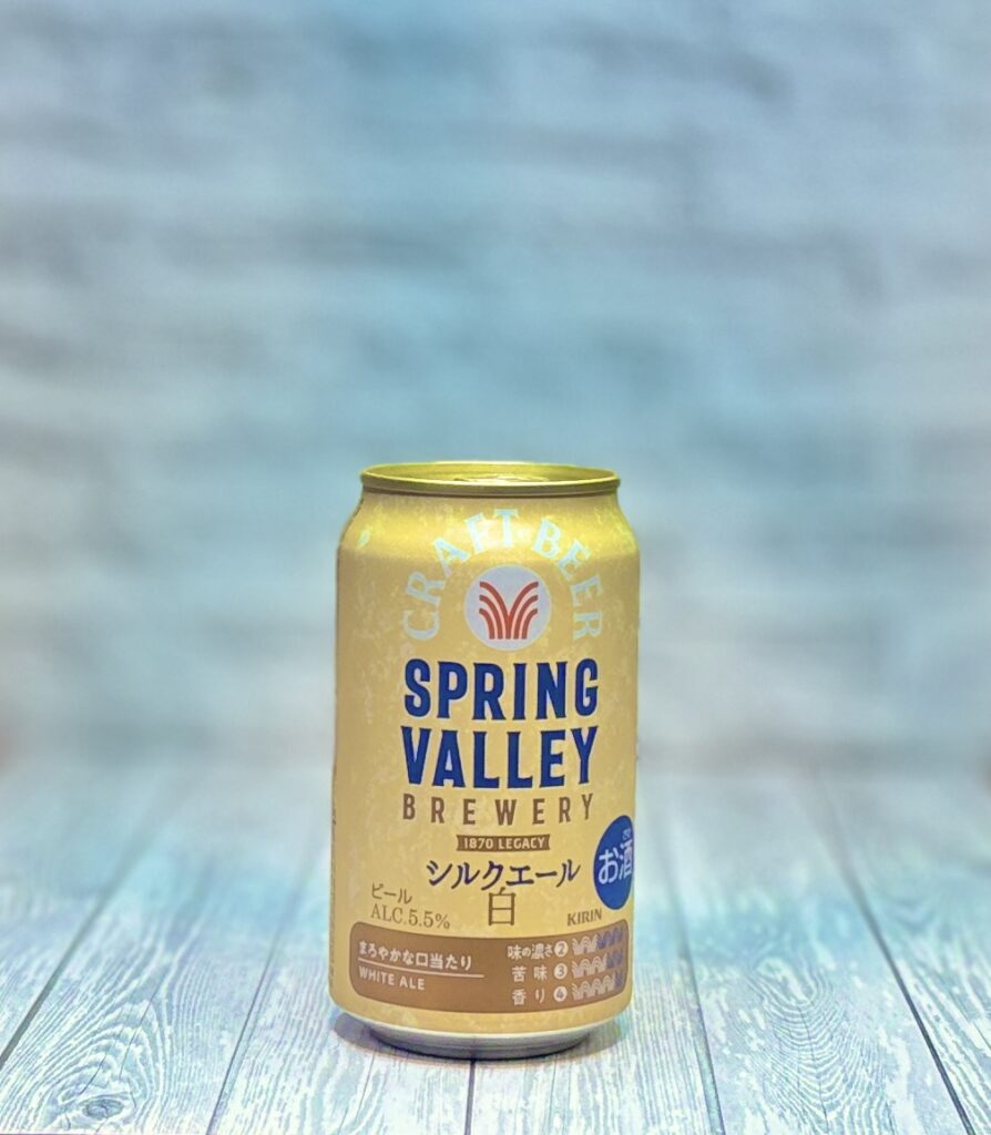 SPRING VALLEY BREWERYのシルクエール白の缶ビールを、淡いブルーの背景と木目調の台の上で撮影した商品写真。やわらかな光の中でゴールドの缶が映え、上品で軽やかな印象があり、食事に合わせるクラフトビールとしての特別感が伝わる一枚。手羽先の塩焼きや和風の副菜と合わせる晩酌シーンにもぴったりで、食中酒や家飲みの楽しさを演出できる。クラフトビール、スプリングバレー、シルクエール白、晩酌のお供、料理に合うビールの紹介用画像として使いやすい写真。