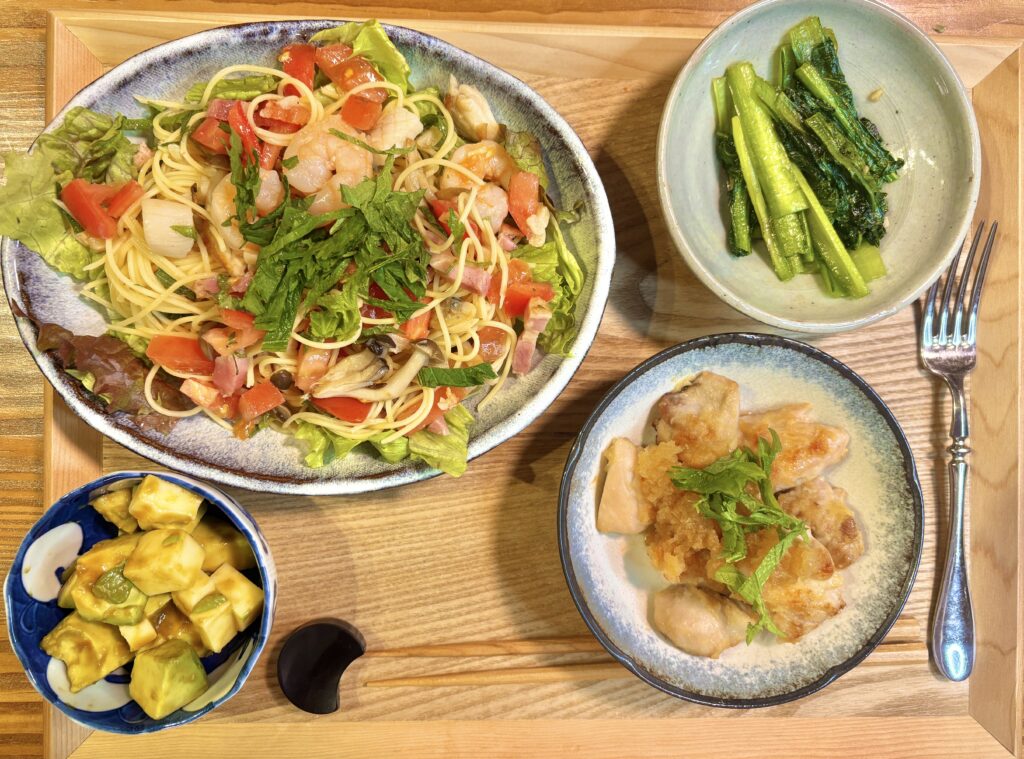 旦那キッチン特製の初夏の献立全体写真。中央にシーフードミックスと貝柱とベーコンのガーリック炒めを混ぜ込んだ冷製サラスパサラダ、右上に小松菜のガーリックソテー、右下に鶏もも肉の生姜みぞれ焼き青紫蘇乗せ、左下にアボカドとモッツァレラチーズのワサビ醤油和えを木製トレーに盛り付けた、白ワインに合う初夏の晩ごはん献立。